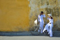 Dos mujeres en ropa tradicional de pie con bicicleta en la calle y hablando, Hoi An, Vietnam - foto de stock