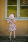 Visão traseira de uma menina vestindo vestido de verão padronizado e chapéu olhando através da janela — Fotografia de Stock