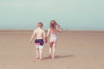 Задній вид дівчинка і хлопчик, тримаючись за руки і ходити на піщаному пляжі — стокове фото
