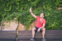Porträt eines Mannes, der auf einer Bank sitzt und ein Selfie mit dem Smartphone macht — Stockfoto