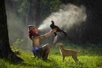 Hombre sosteniendo gallo pájaro y perro de pie sobre hierba verde, Asia - foto de stock