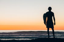 Silhouette dell'uomo in piedi sulla spiaggia al tramonto tenendo tavola da surf — Foto stock