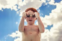 Ingwerjunge fixiert Schwimmbrille vor wolkenverhangenem Himmel — Stockfoto