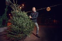 Hipster Hombre sosteniendo un árbol de Navidad por la noche - foto de stock