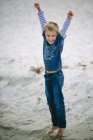 Glücklicher Junge steht mit erhobenen Armen am Sandstrand — Stockfoto