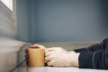 Man holding orange mug of coffee while sitting near window — Stock Photo