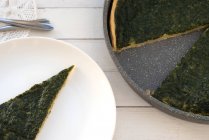 Overhead view of Spinach quiche and slice on white plate over wooden table — Stock Photo