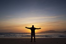 Silueta de hombre de pie con los brazos extendidos en la playa al atardecer, Tarifa, Andalucía, España - foto de stock