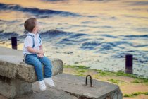 Мальчик в белой рубашке и джинсах сидит на каменной стене в море — стоковое фото