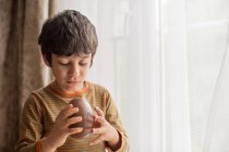Хлопчик стоїть біля вікна і їсть велике шоколадне пасхальне яйце — стокове фото