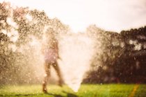 Девушка бежит через разбрызгиватель воды в саду — стоковое фото