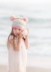 Девушка, стоящая на пляже в смешной шляпе и слушающая ракушку — стоковое фото