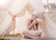 Girl sitting in wigwam on the beach and listening to a seashell — Stock Photo