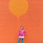 Chica de pie contra una pared naranja pintada con un globo - foto de stock