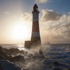 Farol de cabeça de praia contra o nascer do sol com ondas salpicando contra rochas em primeiro plano, East Sussex, Reino Unido — Fotografia de Stock