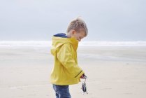Side view of beautiful little boy in yellow raincoat on beach holding mussels — Stock Photo