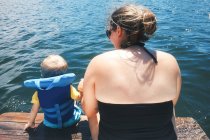 Вид ззаду матері, сидить з сином у надувній куртці на пірсі у воді — стокове фото