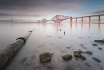 Forth Rail Bridge visto desde el otro lado del mar tranquilo con tubería que corre bajo el agua en primer plano, Queensferry, Escocia, Reino Unido - foto de stock