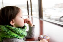 Pensativo chica con donut mirando a través de ventana - foto de stock