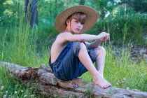 Young boy wearing overalls sitting on tree wearing straw hat — Stock Photo