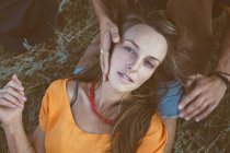 Portrait of young woman lying on grass with male hands on face and looking at camera — Stock Photo