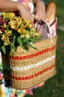 Imagen recortada de la cesta de picnic con flores sostenidas por la mujer en vestido floral - foto de stock