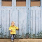 Mignon petit garçon souriant en imperméable jaune debout à l'extérieur des portes verrouillées — Photo de stock