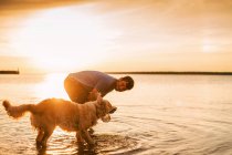 Homem brincando com seu cão golden retriever no lago ao pôr do sol — Fotografia de Stock