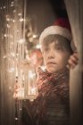 Garçon par une fenêtre portant Noël santa chapeau tenant une lanterne — Photo de stock