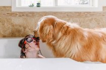 Girl wearing swimming goggles in the bath with her golden retriever dog — Stock Photo