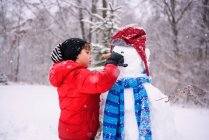 Niño construyendo un muñeco de nieve en el bosque de invierno - foto de stock