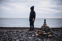 Niño parado en la playa junto a una pila de guijarros, Irlanda - foto de stock