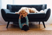 Девочка лежит под диваном и играет со своей собакой — стоковое фото