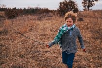 Niño corriendo por el paisaje rural sosteniendo un palo - foto de stock