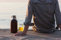 Homem sentado na doca junto à praia com uma cerveja — Fotografia de Stock