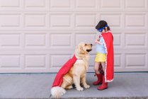 Chica vestida como un superhéroe de pie junto al garaje con su perro golden retriever - foto de stock