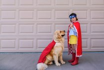 Девушка, одетая как супергерой, стоящая у гаража со своей золотой собакой-ретривером — стоковое фото
