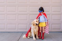Chica vestida como un superhéroe de pie junto al garaje con su perro golden retriever - foto de stock