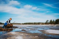 Menino em pé junto a um lago congelado segurando um pau — Fotografia de Stock