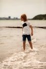 Ragazzo in piedi sulla spiaggia nel surf guardando verso il mare — Foto stock