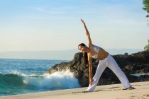 Eine junge Asiatin macht Yoga an einem weißen Sandstrand auf Bali. Sie trägt eine lange weiße Hose und ein Bikini-Oberteil. — Stockfoto