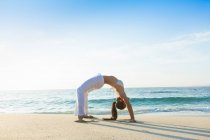 Una joven asiática está haciendo yoga en una playa de arena blanca en Bali. Lleva pantalones blancos largos y un top de bikini.. - foto de stock