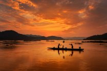 Siluetas de pescadores y embarcaciones en redes fluviales en agua de mar durante la puesta del sol, Tailandia - foto de stock