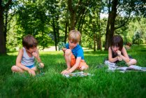 Троє маленьких дітей сидять на задньому дворі читають книги та їдять свіжі овочі — стокове фото