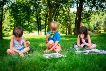Três crianças pequenas sentadas no quintal lendo livros e comendo vegetais frescos — Fotografia de Stock