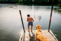 Young boy thinking about leaping off a dock into a lake with a dog - foto de stock