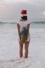 Mujer con un sombrero de Santa Navidad de pie en el océano surf sosteniendo una piña a sus espaldas, Haleiwa, Hawai, América, EE.UU. - foto de stock