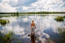 Молодий хлопчик впадає в мирне озеро з відображенням неба і хмар з лопатою — стокове фото