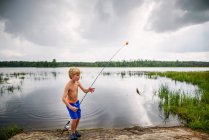 Young boy holding fish by peaceful lake with reflection of sky and clouds — Stock Photo