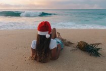 Mulher vestindo um chapéu de Natal Santa deitado na praia ao lado de um abacaxi, Haleiwa, Havaí, América, EUA — Fotografia de Stock
